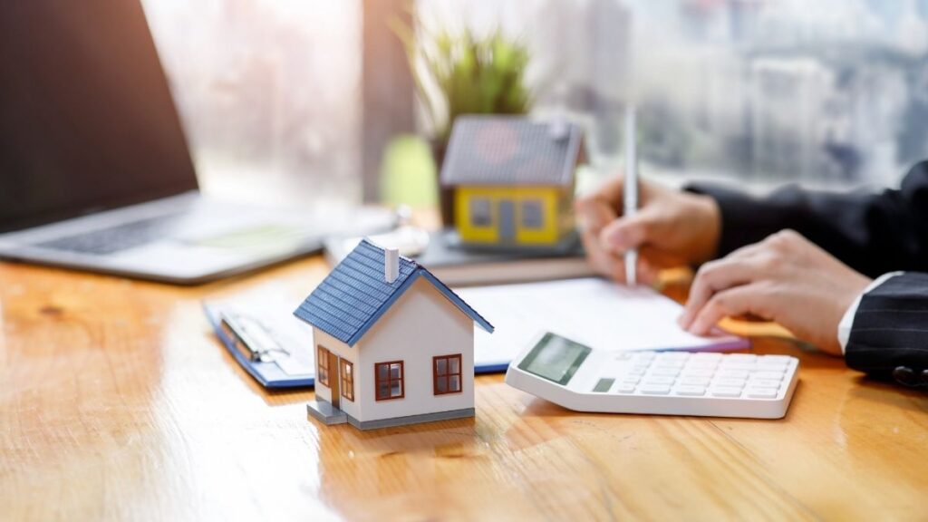 Un homme rédige sur une feuille, avec devant lui une maquette de maison et une calculatrice