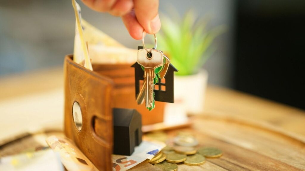 Une main tenant un trousseau de clés au-dessus d’un porte-monnaie ouvert, avec des billets et des pièces posés sur une table